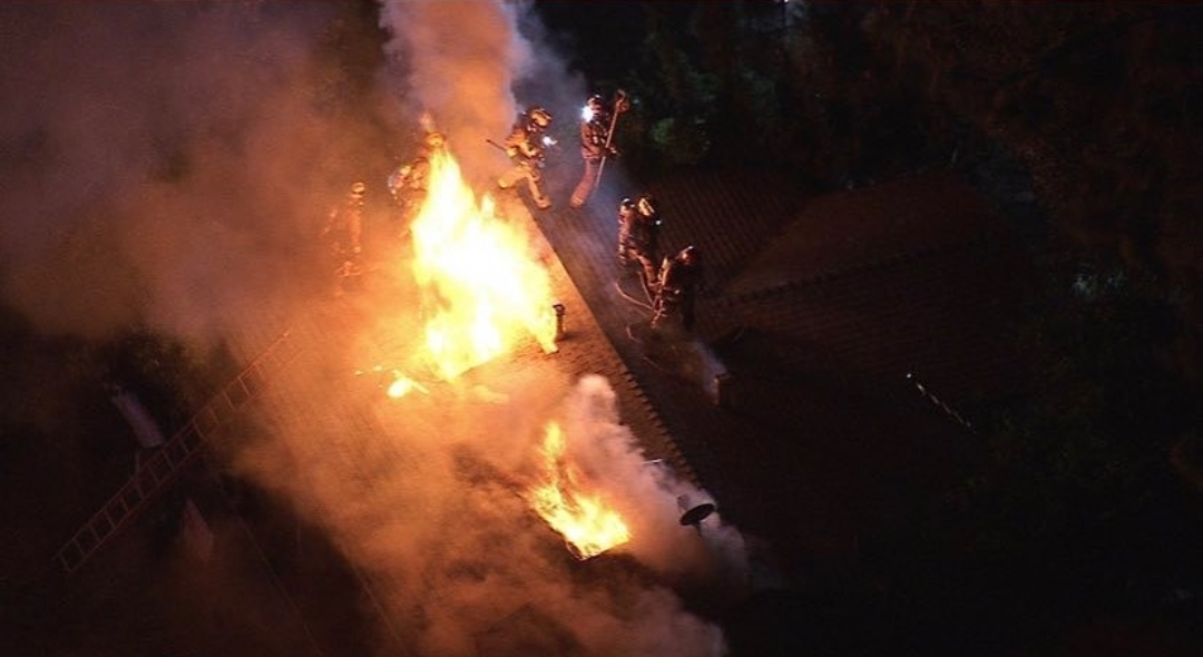 Photo of firefighters standing atop a building with flames coming out of the roof